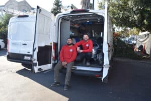Two men in red shirts sit at the open back of a white HVAC van parked outdoors on a sunny day.