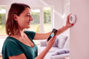 A smiling woman using her phone while adjusting a thermostat in a home.