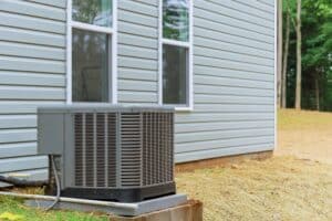 A central air conditioning unit is installed outside next to a light gray house with two windows visible.