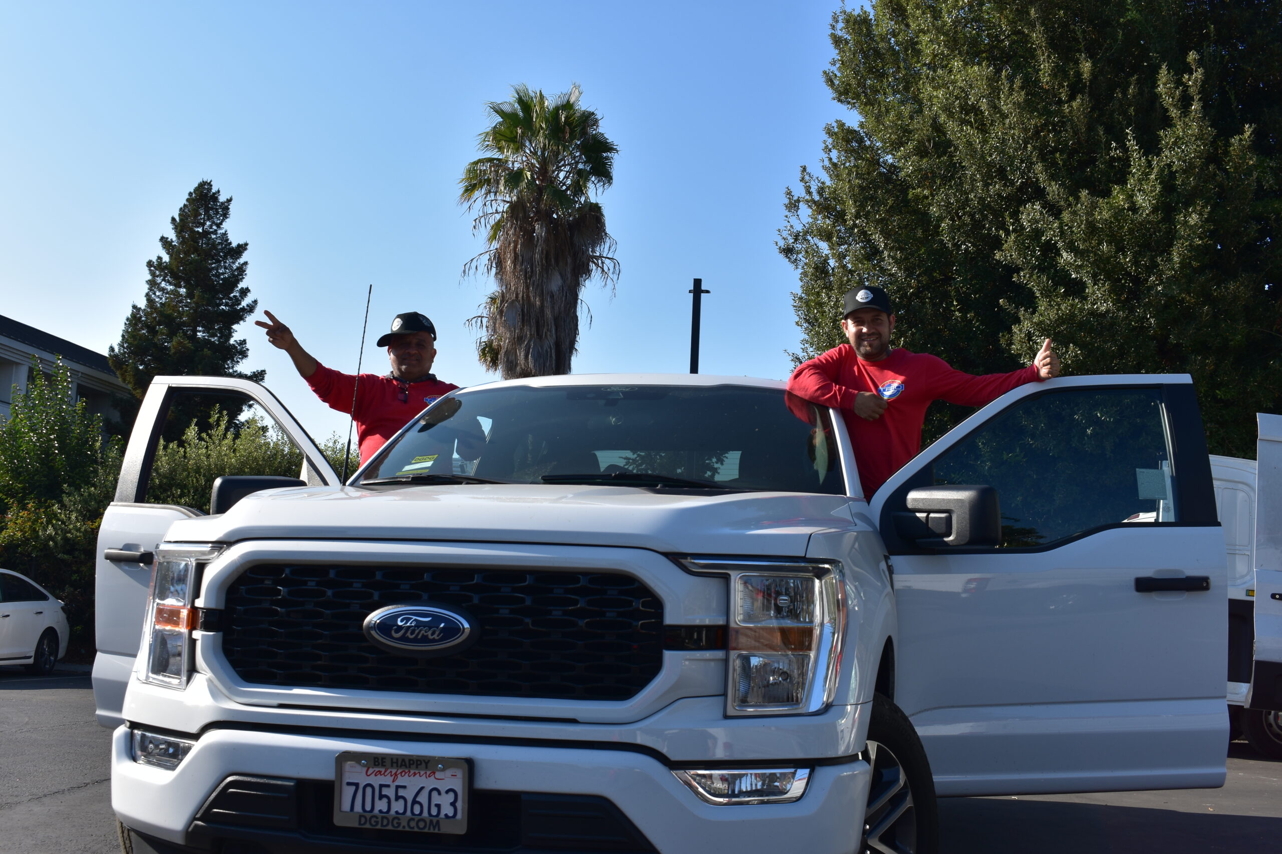 Two men in red shirts posing happily with a white Ford truck, open doors, palm and trees in background.