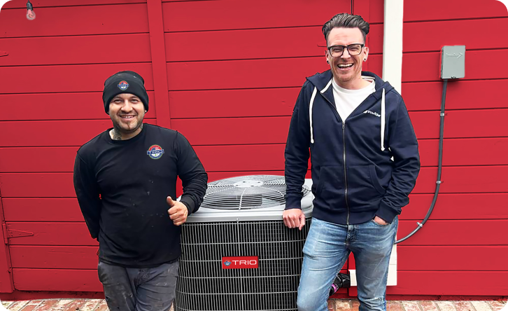 Two men standing by a TRIO air conditioning unit outside a red building, both smiling at the camera.