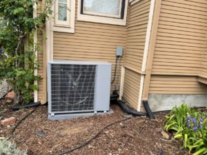 A gray outdoor air conditioning unit is installed next to the beige siding of a house; black flexible conduit tubes run along the exterior wall, and ground cover mulch surrounds the heating and cooling area.