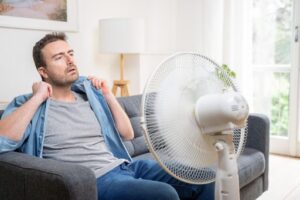 A man sitting in a hot room on a couch in front of a fan.