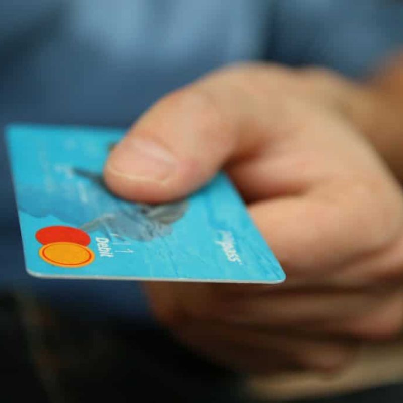 A person holds out a blue debit card with the Mastercard logo visible, offering it forward—perfect for paying your next heating or plumbing bill.