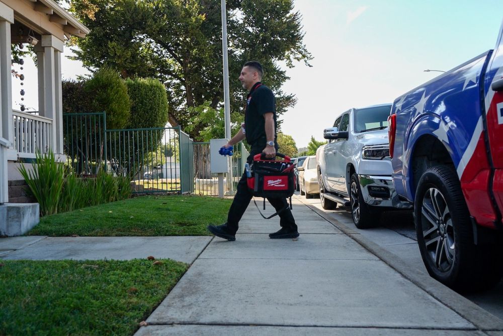 Technician walking with a tool bag towards a house, passing parked service trucks on a sunny day.
