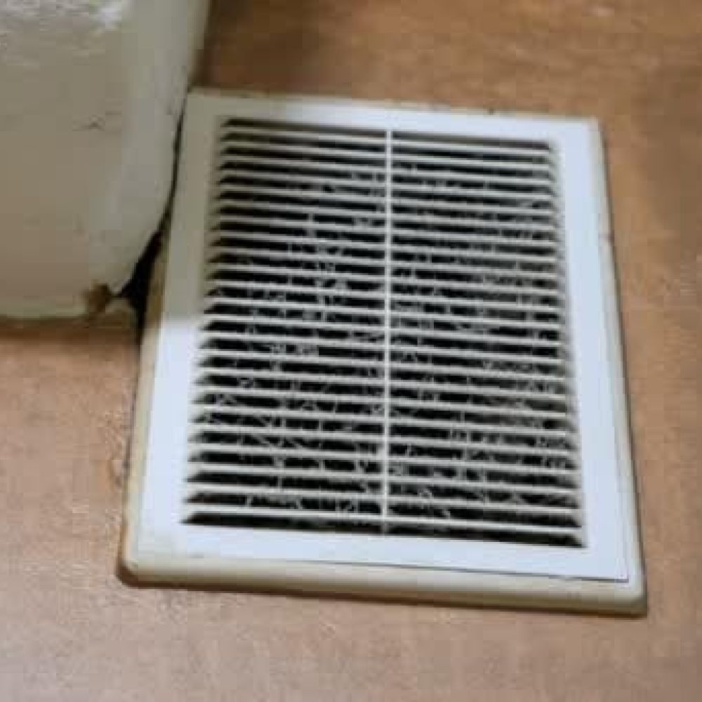 Dirty air vent on a brown floor near a wall corner, highlighting the need for cleaning and maintenance in home environments.
