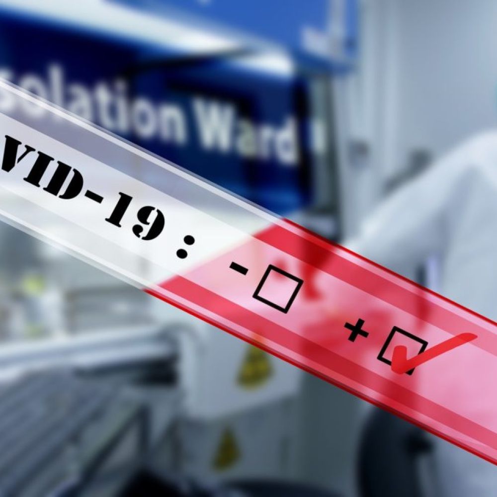 A COVID-19 test tube shows a positive result, with a healthcare worker and an "Isolation Ward" sign visible in the background, highlighting the importance of proper HVAC systems in healthcare settings.