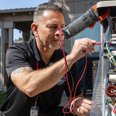 Technician repairing an outdoor HVAC unit using tools and electrical test equipment in a residential backyard.