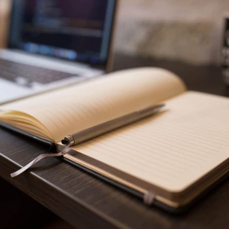 An open lined notebook with a pen rests on a desk next to a laptop and a mug, perfect for jotting down heating and air conditioning notes.