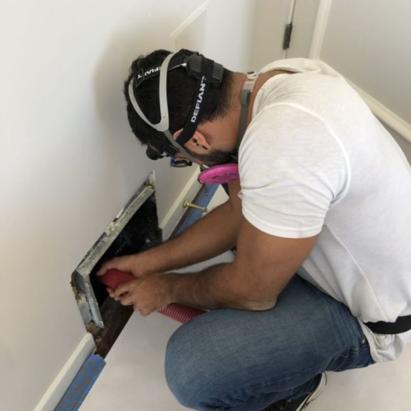 Man cleaning air duct with hose, wearing protective gear in residential setting. Home maintenance and safety concept.