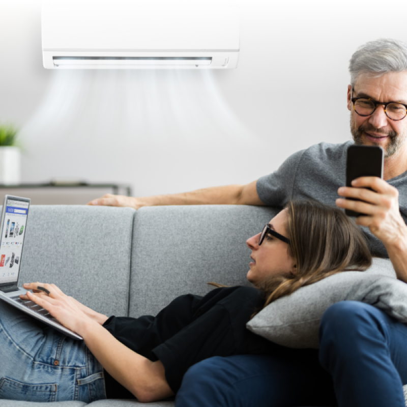 Couple relaxing on sofa with laptop and smartphone, enjoying air-conditioned comfort at home.
