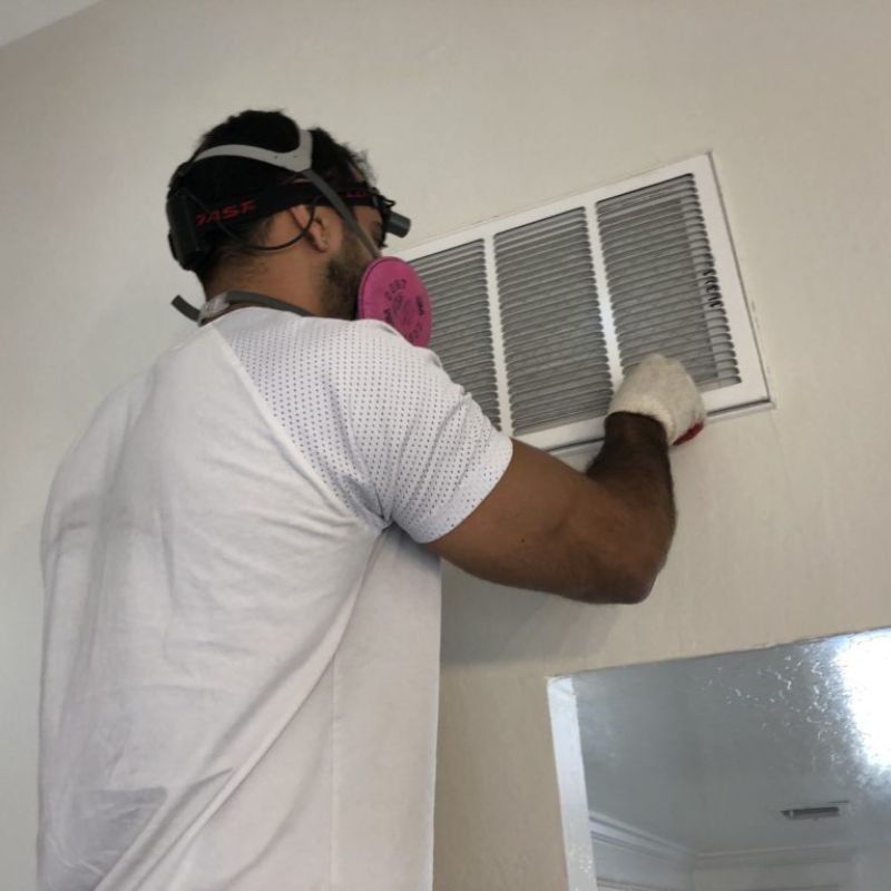 A person wearing safety glasses, gloves, and a respirator mask is cleaning or inspecting an air vent on an interior wall, ensuring the air conditioning system remains safe and efficient.
