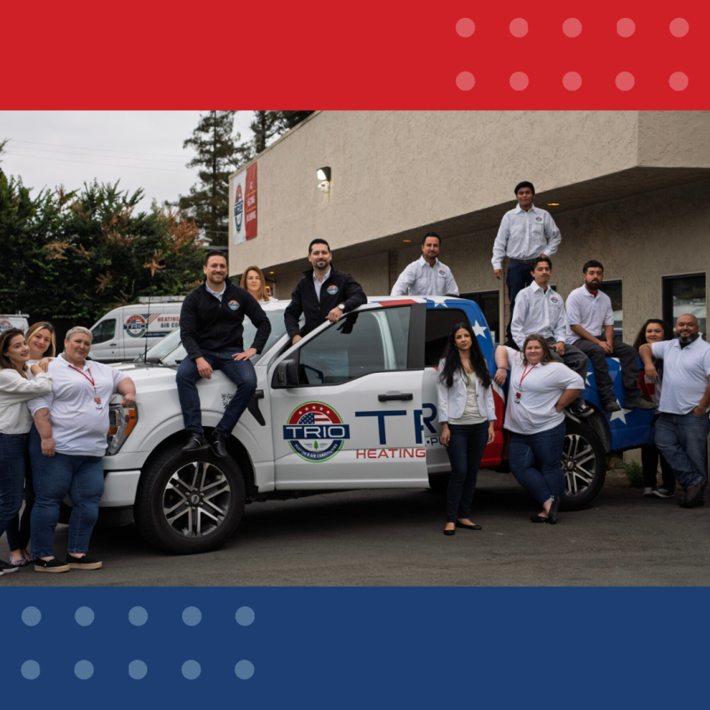 A group of eleven people poses around and on top of a Trio Heating & Air truck, showcasing their hvac expertise outside a commercial building.