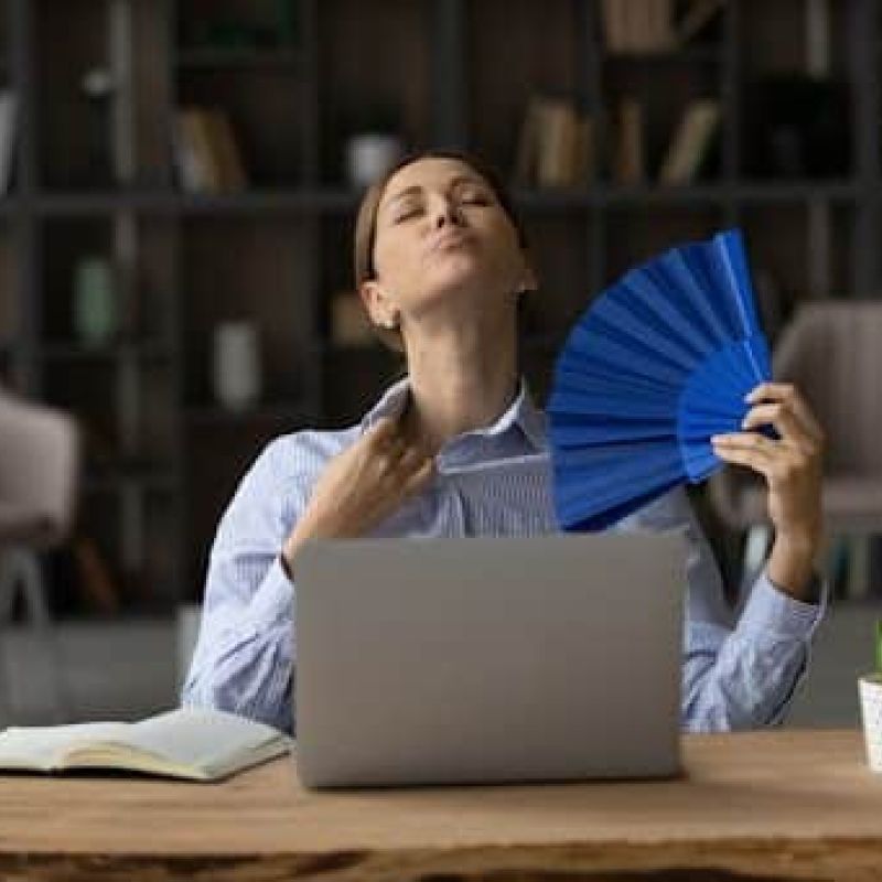 Woman fanning herself while sitting at desk in home office because AC is blowing hot air.