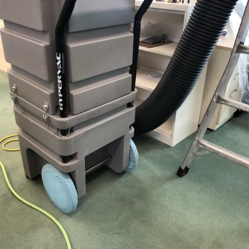 A gray industrial vacuum cleaner with large wheels and a black hose, ideal for hvac or air conditioning maintenance, is positioned next to a metal ladder and a white cabinet on a green carpeted floor.