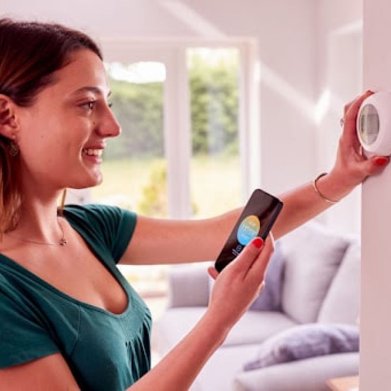 A smiling woman using her phone while adjusting a thermostat in a home.