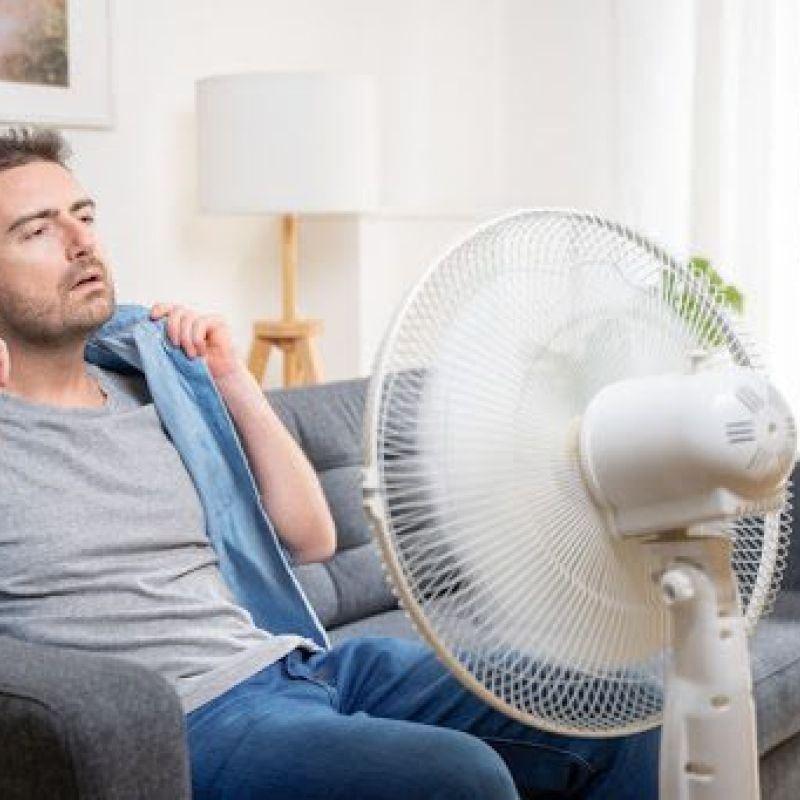 A man sitting in a hot room on a couch in front of a fan.