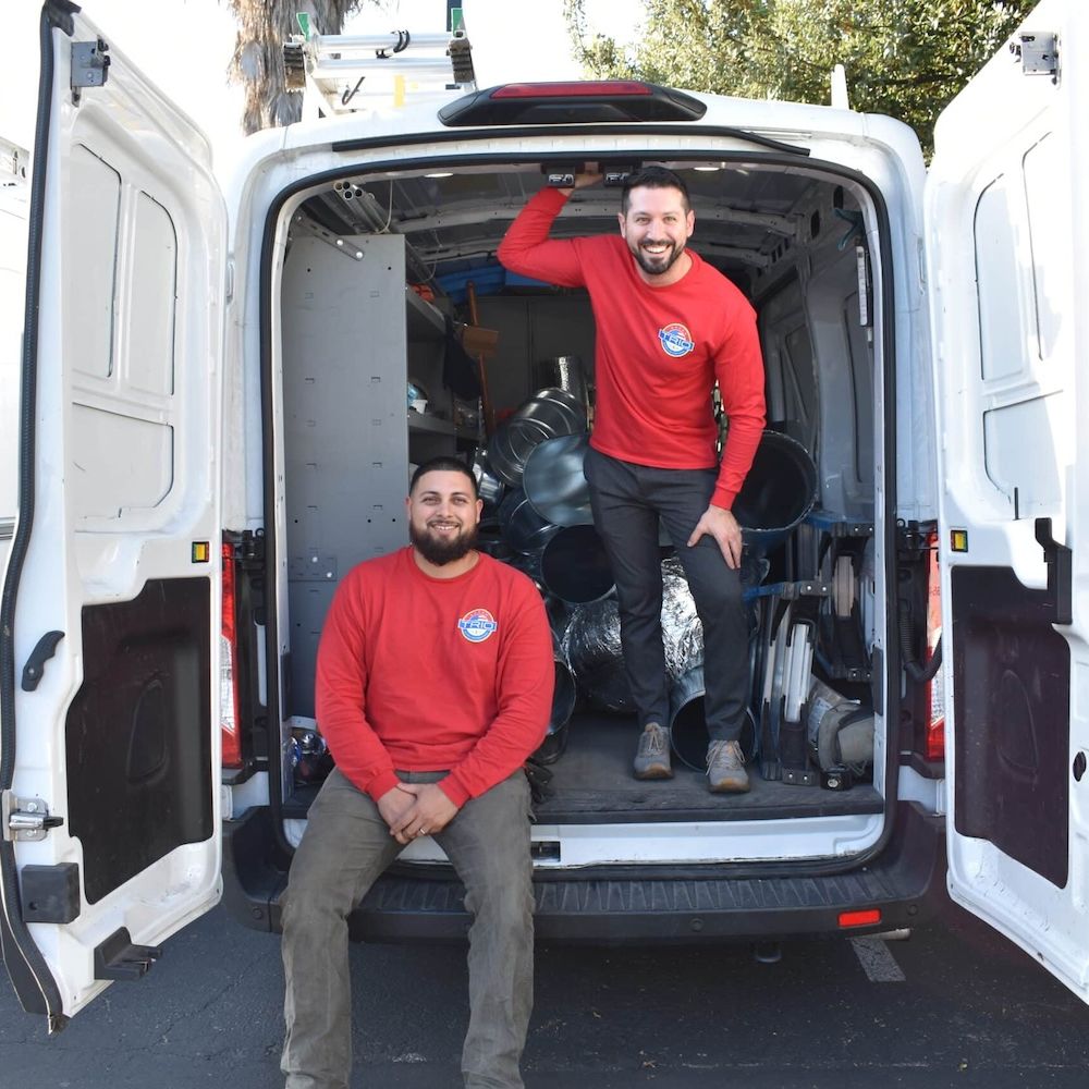 Two smiling workers in red shirts sitting in an open white van filled with equipment.