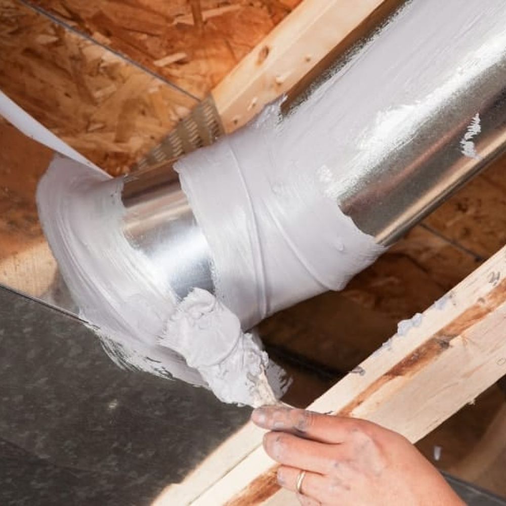 A person applies sealant to a metal HVAC duct joint in a wooden ceiling structure, ensuring the heating system operates efficiently.