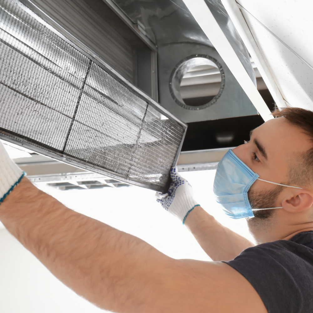 A man wearing gloves and a mask removes a filter from an air duct on the ceiling, likely for cleaning or replacement. A ventilation icon is visible in the lower left corner.