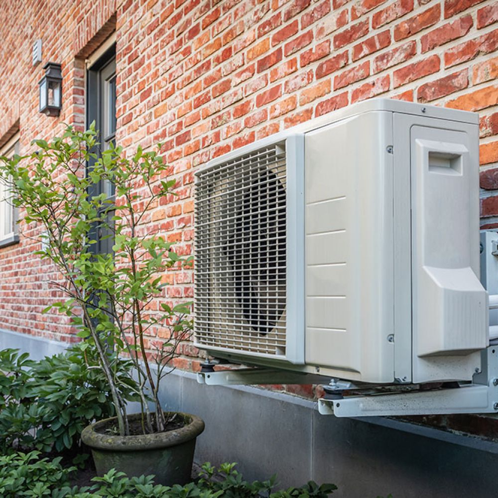 Outdoor air conditioning unit mounted on a brick wall, surrounded by plants for energy-efficient home cooling.
